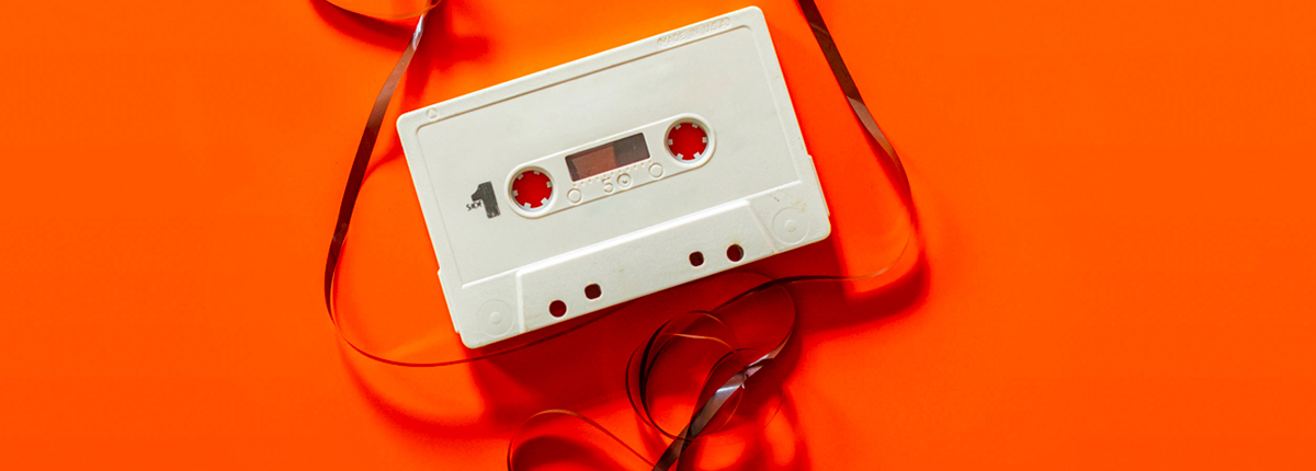 A white cassette tape, with its film pulled out and strung around it, lays atop a bright orange backdrop, invoking a feeling of energetic nostalgia