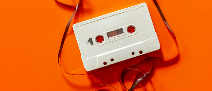 A white cassette tape, with its film pulled out and strung around it, lays atop a bright orange backdrop, invoking a feeling of energetic nostalgia